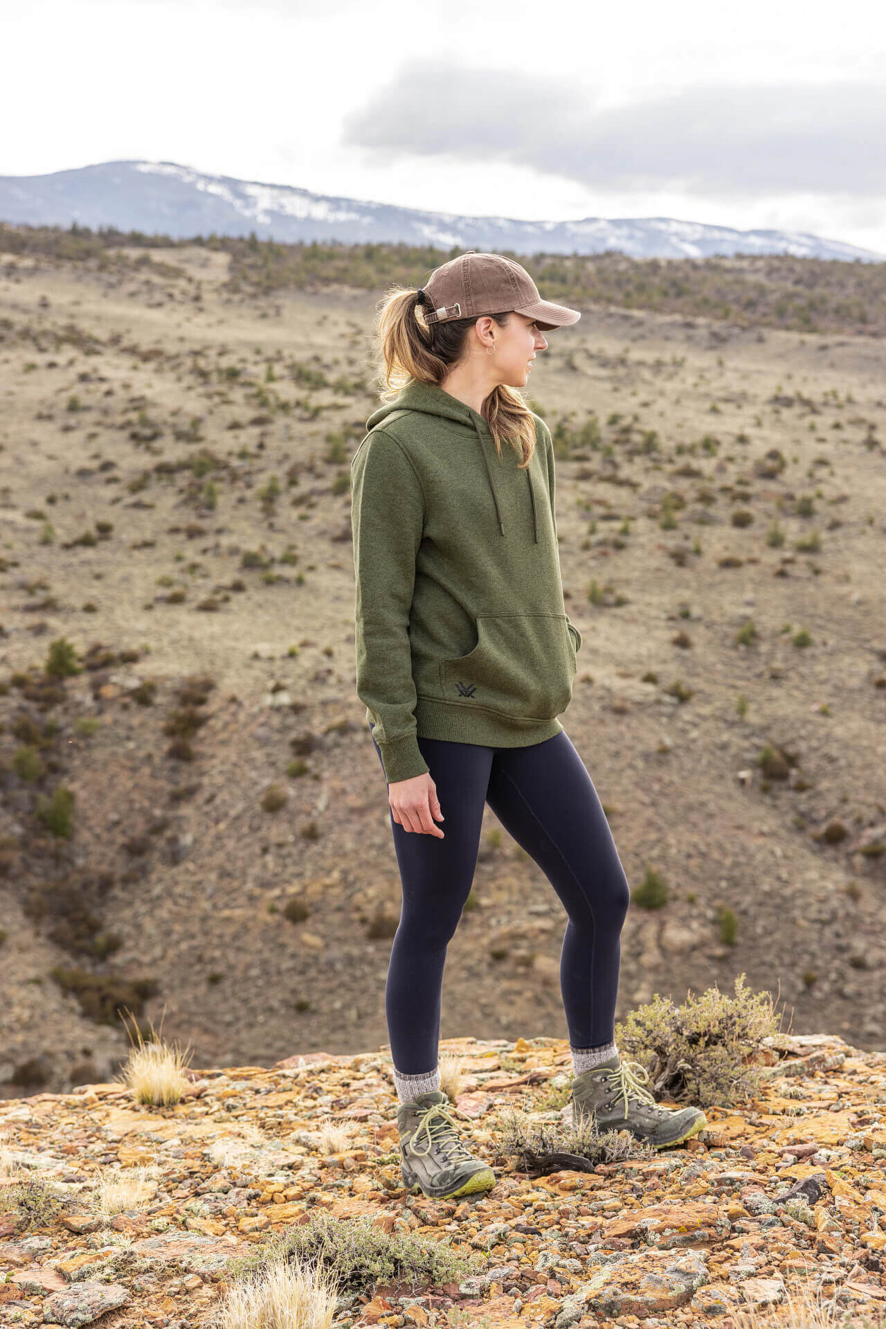 Eine Frau trägt einen grünen Hoodie und eine Kappe, während sie draußen in einer bergigen Landschaft steht.