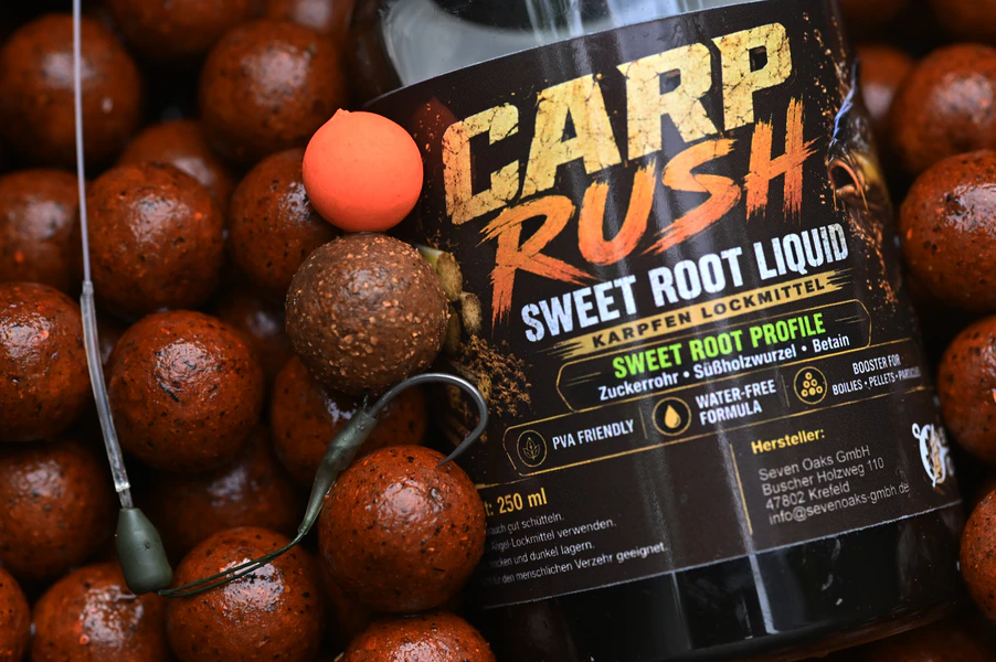 Flasche Carp Rush Sweet Root Liquid Karpfenlockmittel umgeben von braunen Boilies und montiertem Karpfen-Rig