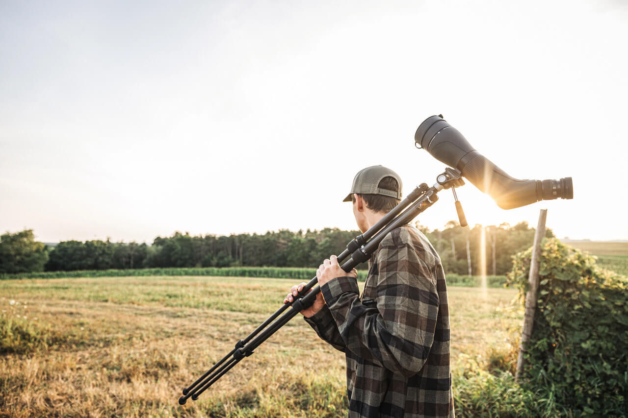 Person trägt ein Stativ mit Fernglas über der Schulter und blickt über ein Feld, Sonnenuntergang im Hintergrund