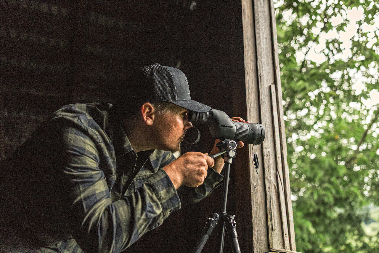 Mann in kariertem Hemd und Baseballkappe schaut durch ein Spektiv auf einem Stativ aus einem Fenster in der Natur