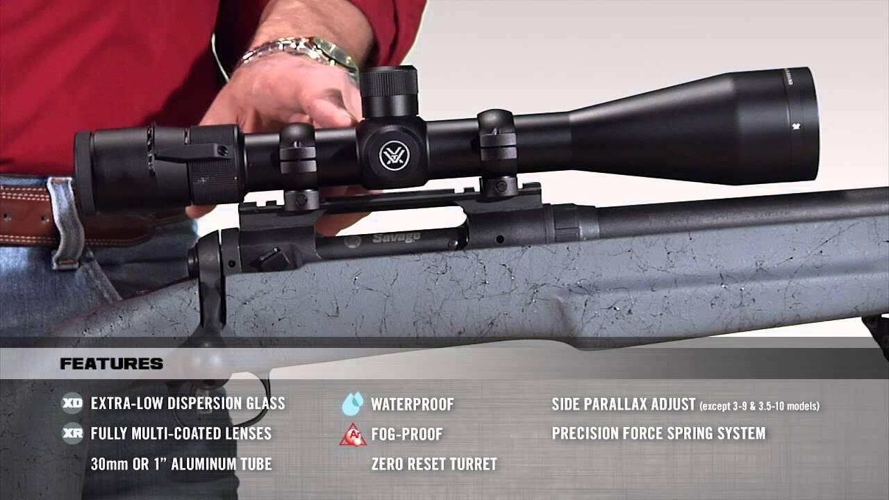Nahaufnahme eines auf einem Gewehr montierten Zielfernrohrs mit Funktionen wie wasserdicht, beschlagfrei und extra-niedriger Dispersion. Eine Hand justiert das Gerät.