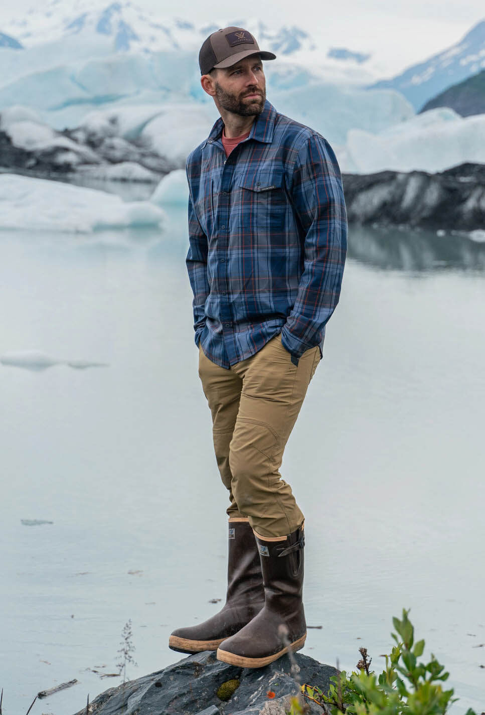 Ein Mann trägt kariertes Hemd und Stiefel, steht auf einem Felsen vor einer beeindruckenden Gletscherlandschaft.