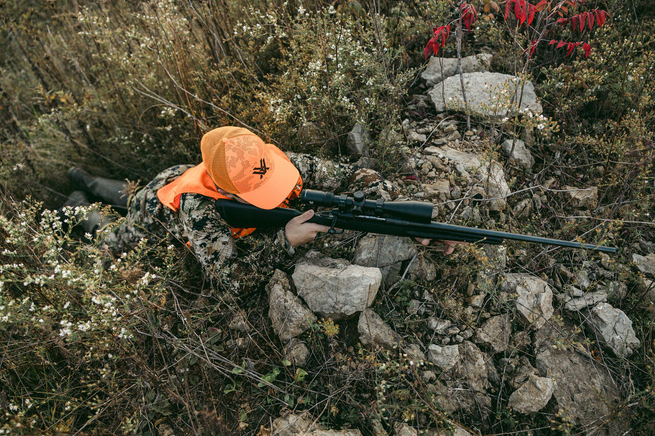 Person in Tarnkleidung und orangefarbener Schirmmütze hält Gewehr und liegt im Gras zwischen Steinen