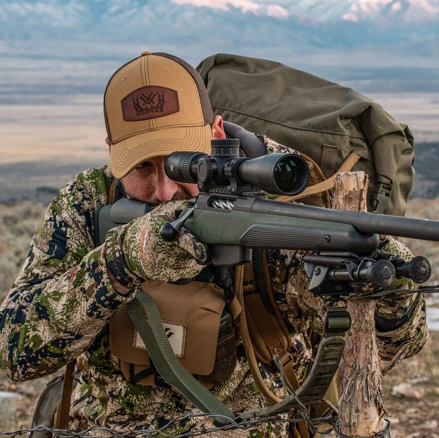 Ein Jäger in Tarnkleidung zielt mit einem Gewehr in einer bergigen Landschaft bei Tageslicht