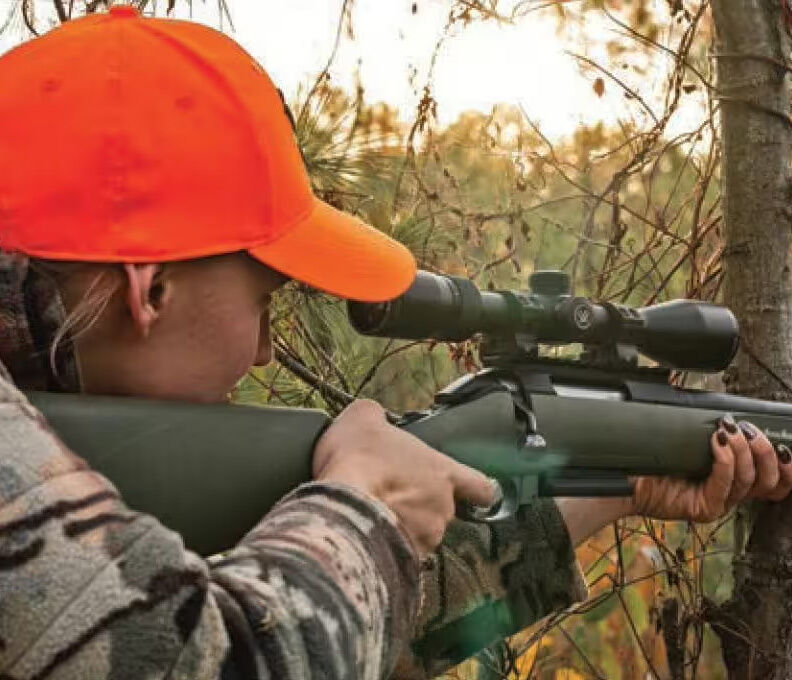 Frau im Tarnanzug mit orangefarbener Mütze zielt mit einem Gewehr im Wald
