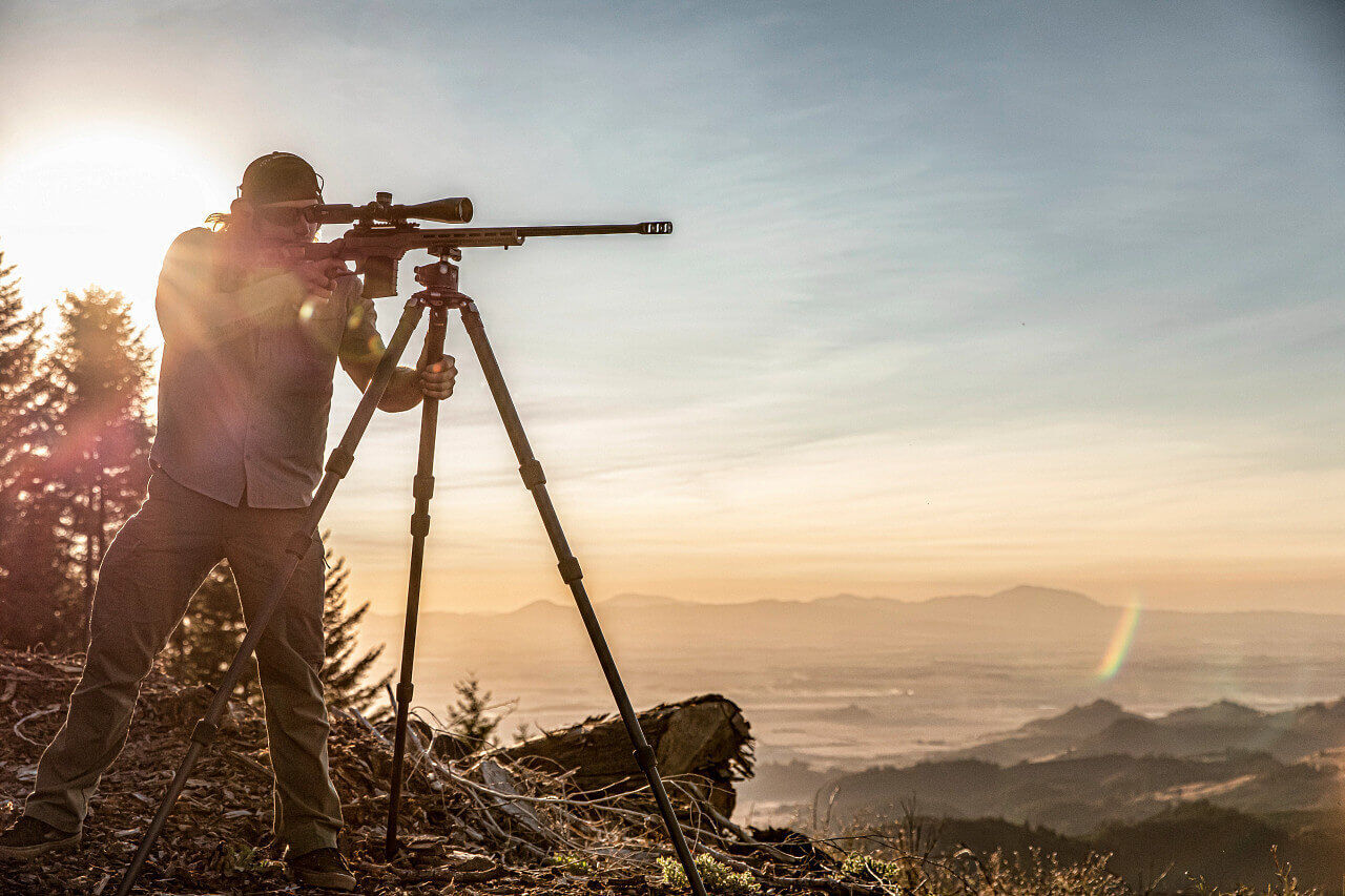 Ein Jäger zielt mit einem Gewehr, das auf einem stabilen Stativ befestigt ist, während die Sonne über einem hügeligen Landschaftspanorama aufgeht.