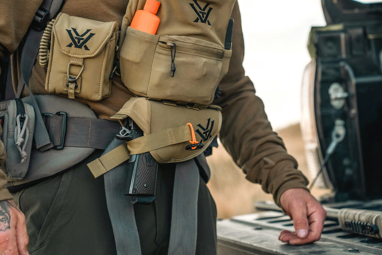 Nahaufnahme einer Person mit taktischer Wanderausrüstung, mehreren Taschen und Gürteltaschen, eine davon mit orangefarbenem Gegenstand bestückt, im Freien stehend.