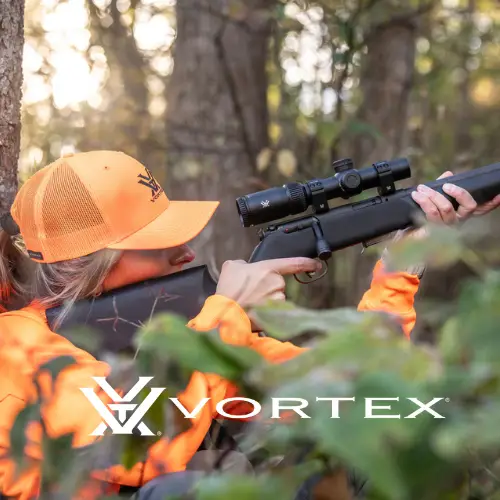 Jägerin in orangefarbener Vortex-Kappe und Jagdbekleidung zielt mit Gewehr und Vortex-Zielfernrohr im dichten Wald