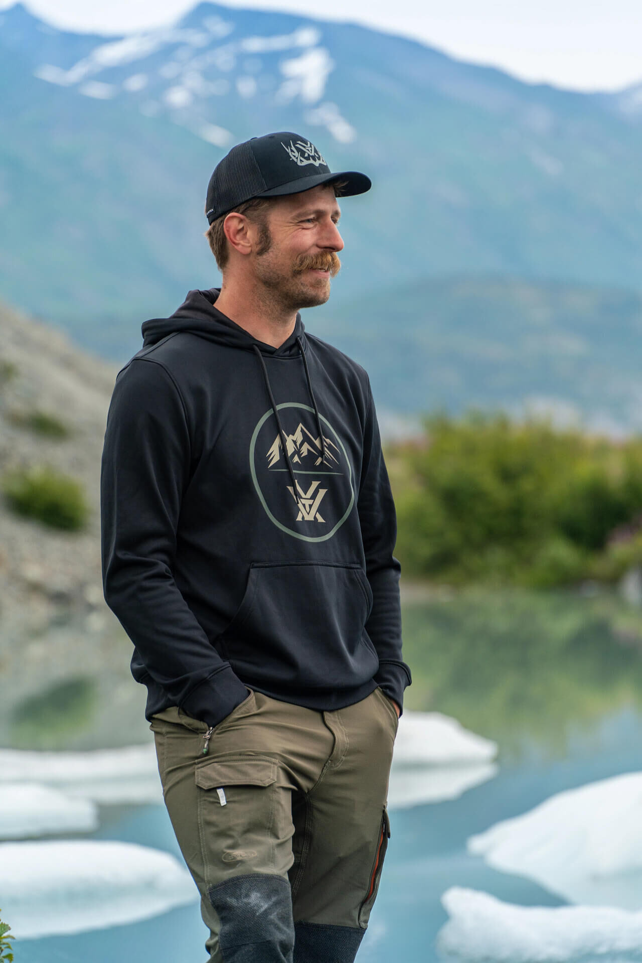Mann mit schwarzem Kapuzenpullover und Cargohose in einer malerischen Berglandschaft