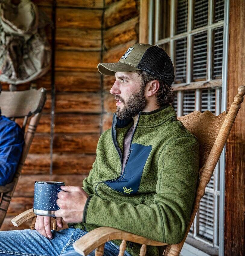 Mann in grüner Jacke und Mütze, sitzt auf einer Holzveranda und hält eine Tasse