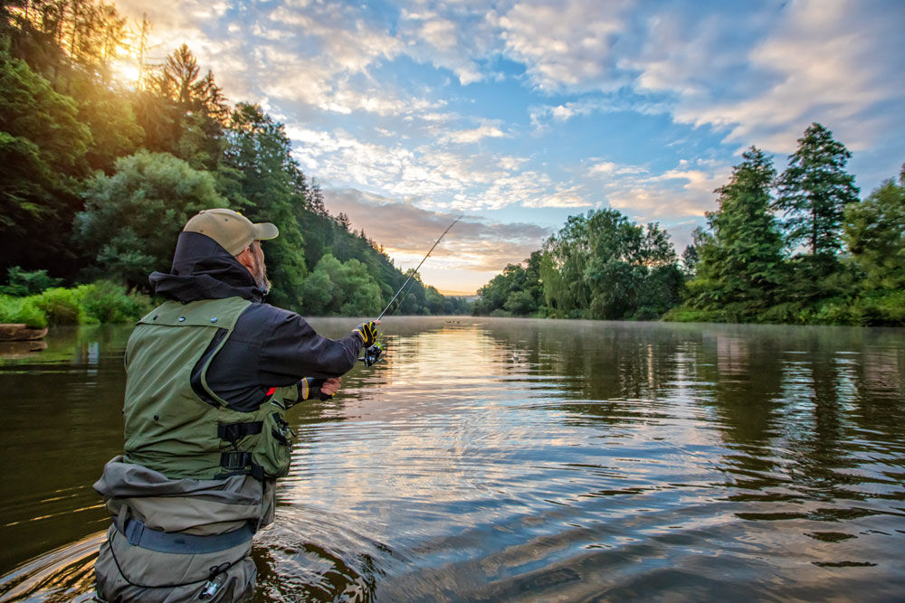 Ein Mann beim Fliegenfischen im Fluss, umgeben von Wald und unter einem klaren, morgendlichen Himmel