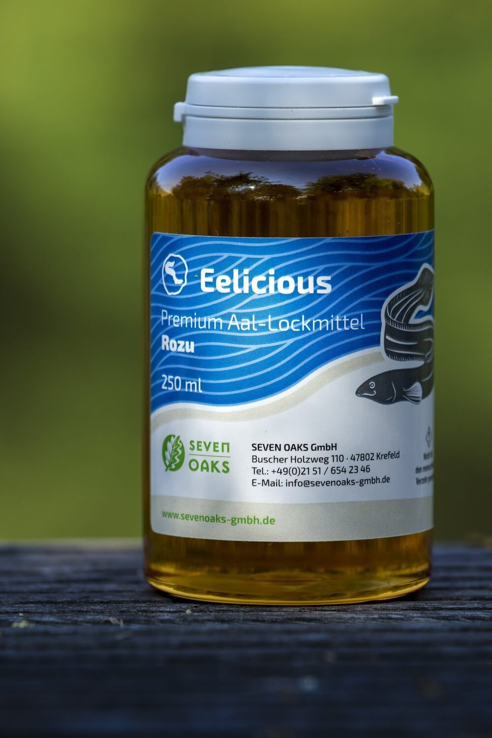 Flasche mit Eelicious Premium Aal-Lockmittel Rozu, 250 ml, von SEVEN OAKS GmbH auf Holztisch vor grünem Hintergrund