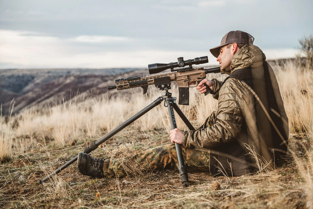 Ein Mann in Outdoor-Kleidung sitzt in einem Feld und zielt mit einem Gewehr auf einem Stativ in die Ferne.