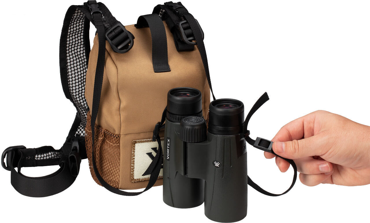 Eine Hand hält ein Fernglas neben einer braunen Tasche mit Tragegurten, geeignet für Naturbeobachtungen.