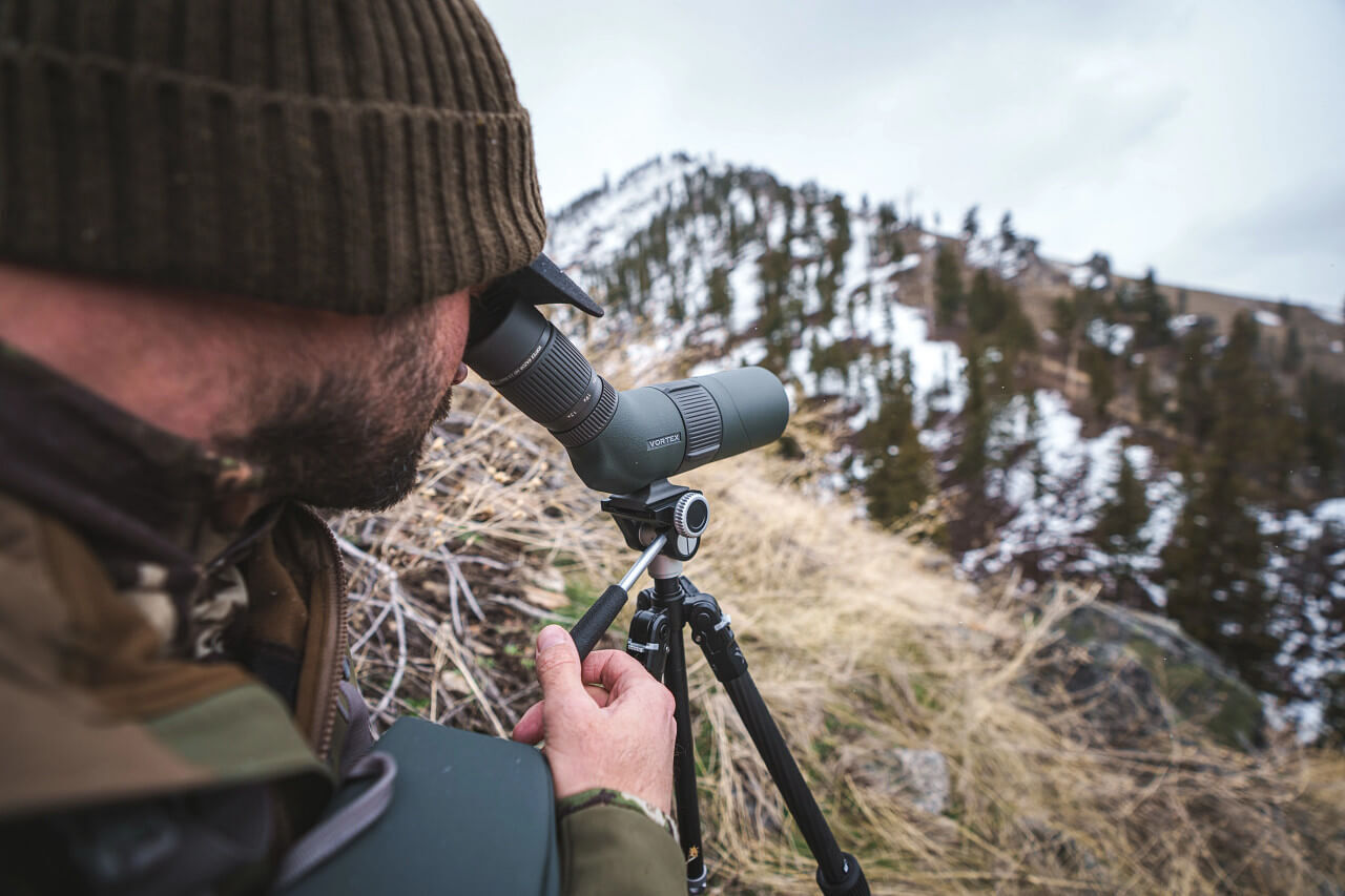 Person betrachtet durch ein Spektiv eine verschneite Berglandschaft