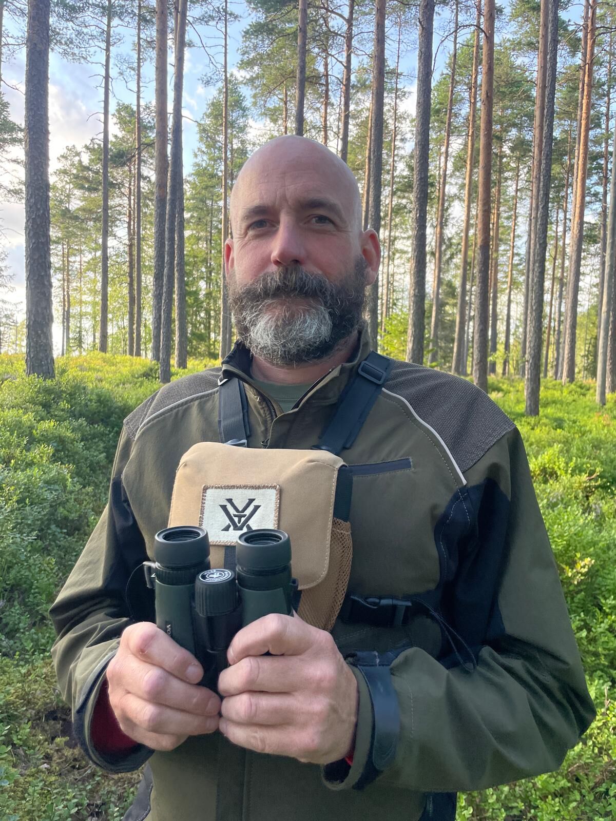 Mann in Outdoor-Jacke hält ein Fernglas vor einer Brusttasche mit Logo und steht in einem lichten Nadelwald