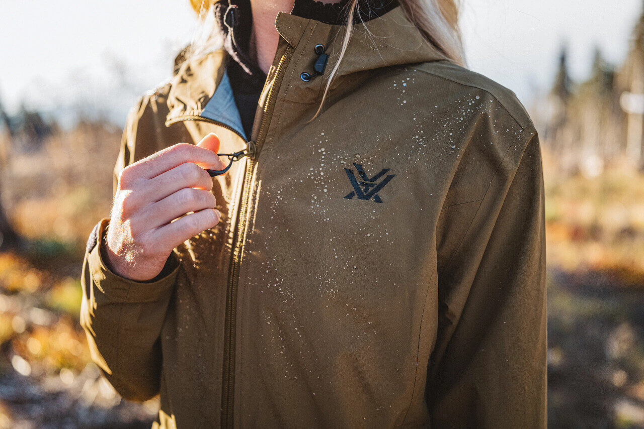 Person trägt eine braune, wasserabweisende Outdoor-Jacke mit Reißverschluss, verziert mit Wassertropfen