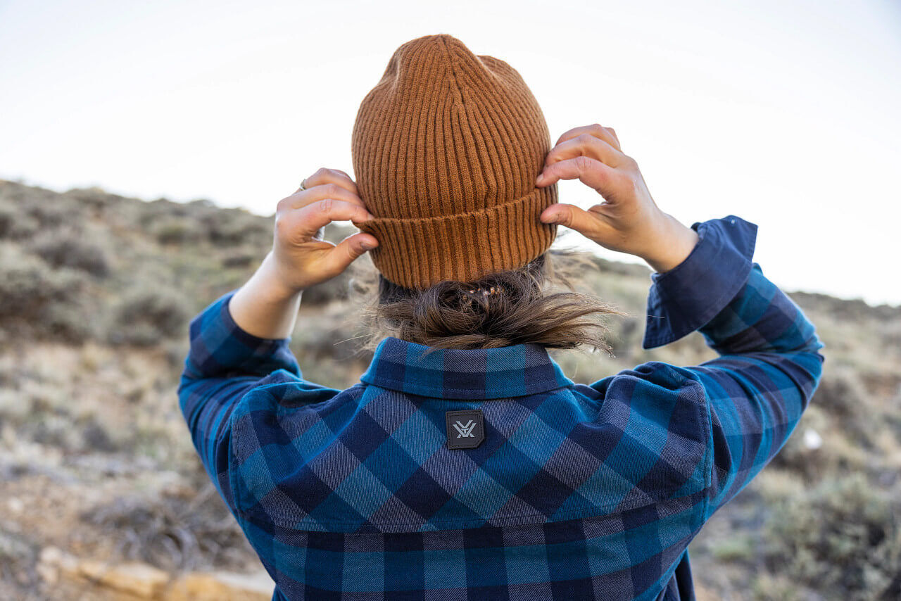 Rückansicht einer Person in blauer Flanelljacke und brauner Strickmütze, die sich in einer natürlichen Umgebung befindet, mit Blick auf eine Landschaft