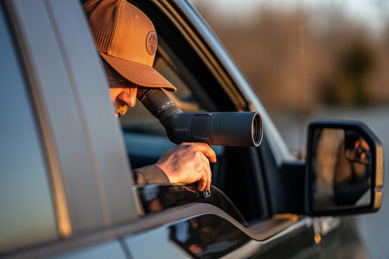 Mann mit Baseballcap beobachtet durch ein Spektiv aus einem Autofenster bei Sonnenuntergang