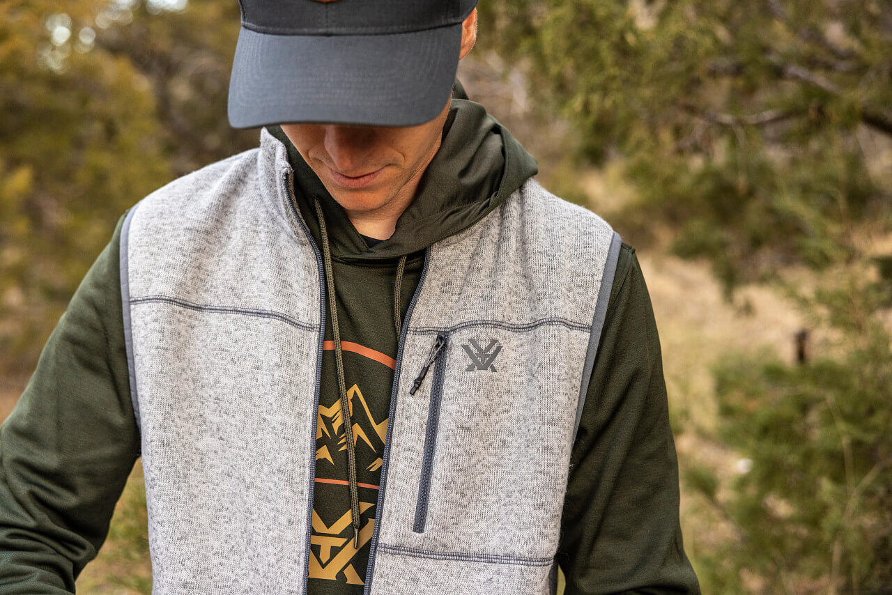 Person trägt graue Outdoor-Weste über einem khakifarbenen Kapuzenpullover im Freien, Kopf von Baseballcap verdeckt