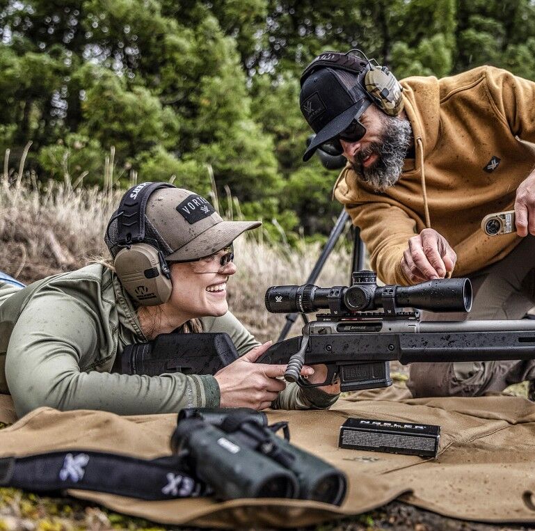 Zwei Personen liegen auf einer Matte im Freien und üben mit einem Gewehr und einem Zielfernrohr das Schießen.