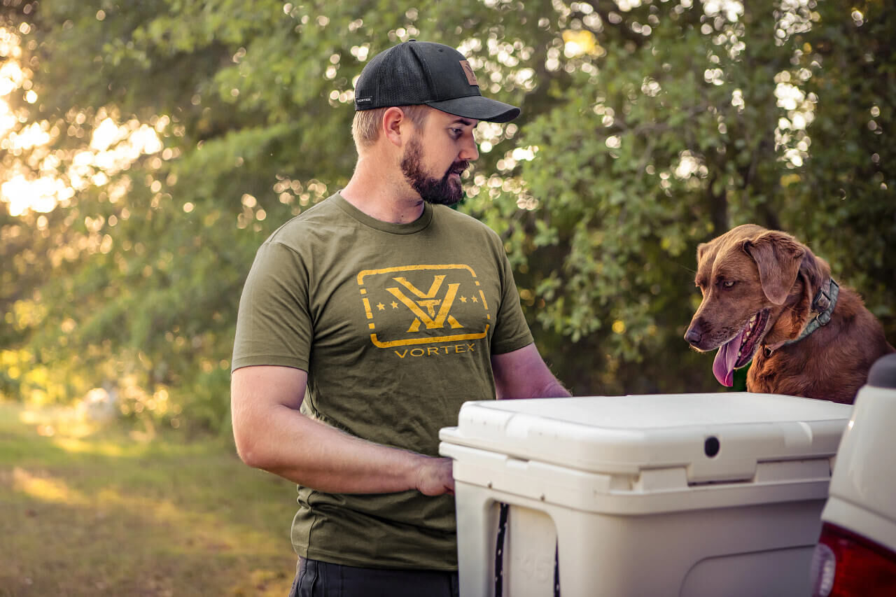 Ein Mann in einem grünen Vortex-T-Shirt steht neben einem Hund und einer Kühlbox im Freien.