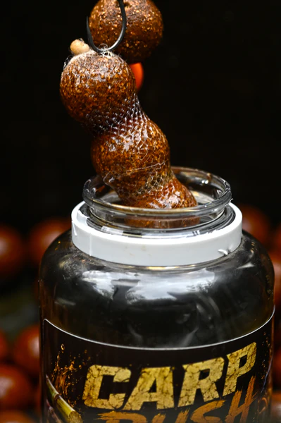 Gefülltes PVA-Netz mit Karpfenködern wird aus einem offenen Glas Seven Oaks CARP RUSH Sweet Root Liquid 250 ml als süß-würziges, PVA-freundliches Karpfen Lockmittel gezogen