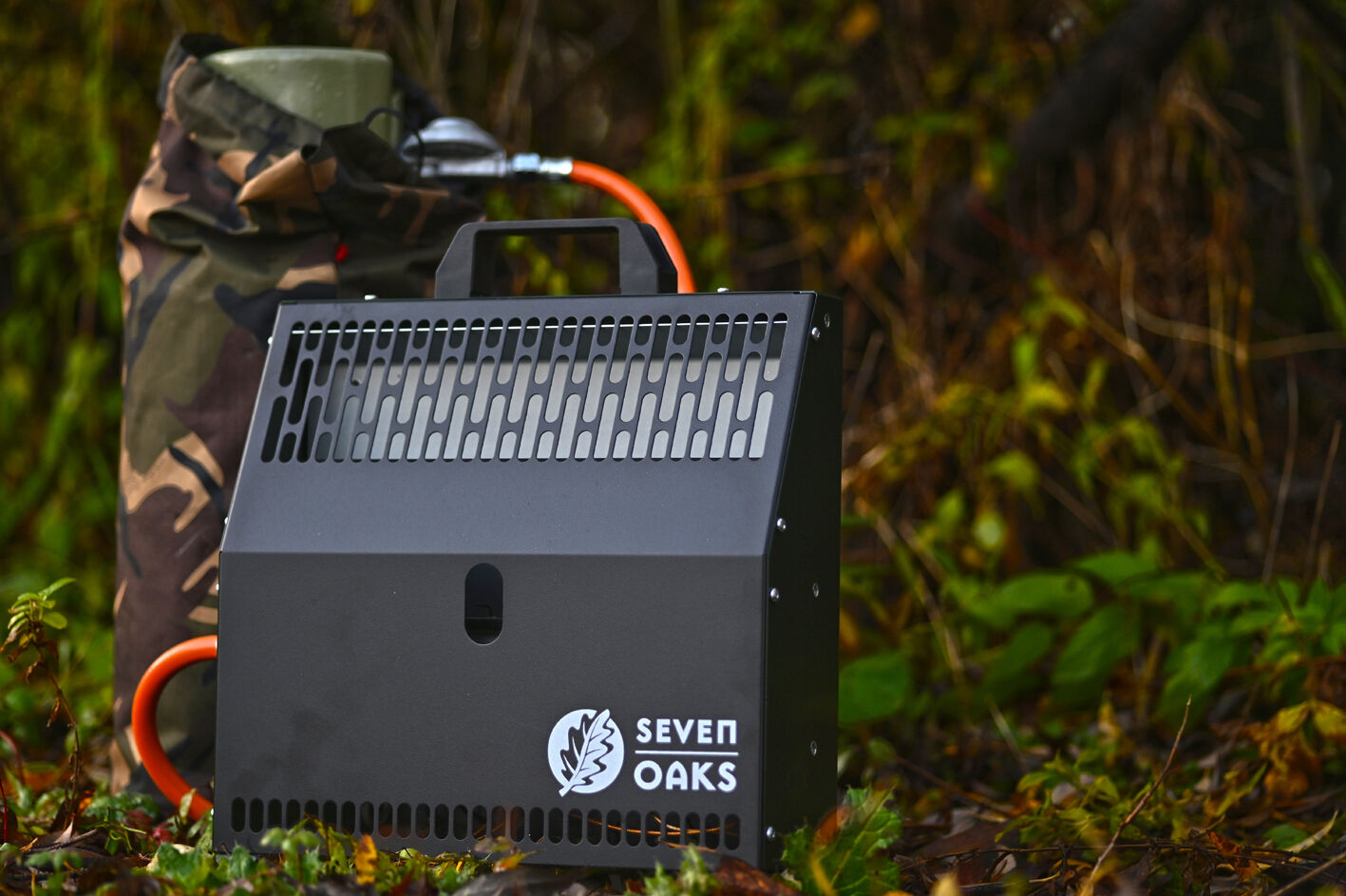 Eine tragbare Heizvorrichtung der Marke Seven Oaks steht neben einer Gaskartusche mit Tarnbezug im Wald.