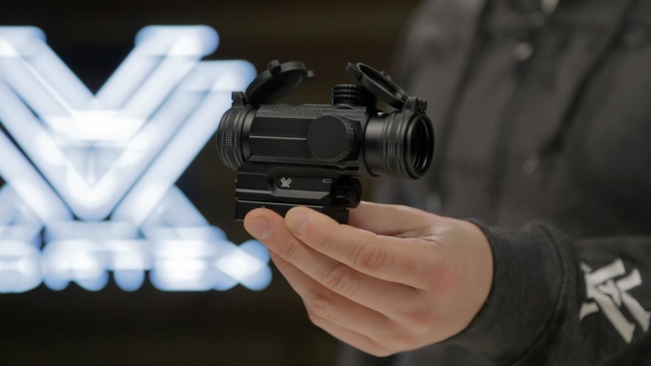 Person hält ein Red Dot Visier mit Flip-Up Objektivkappen vor einem unscharfen Hintergrund