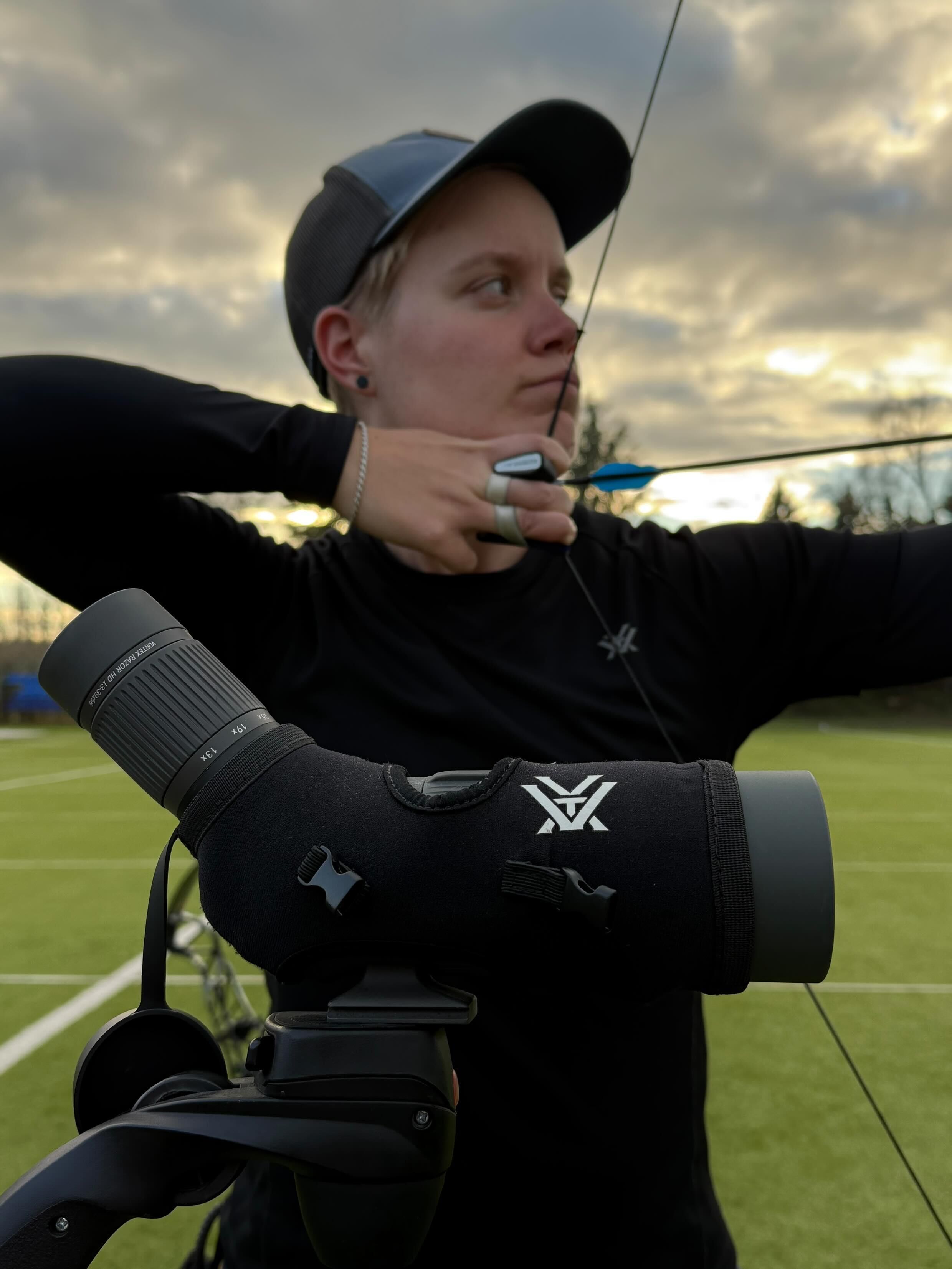 Person im schwarzen Langarmshirt und Baseballkappe zieht einen Bogen, während ein Fernglas im Vordergrund fokussiert ist. Der Himmel ist bewölkt.