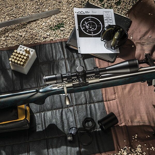 Ein Präzisionsgewehr mit Zielfernrohr, umgeben von Schießscheiben, Gehörschutz und Munition auf einer Schießmatte im Freien.