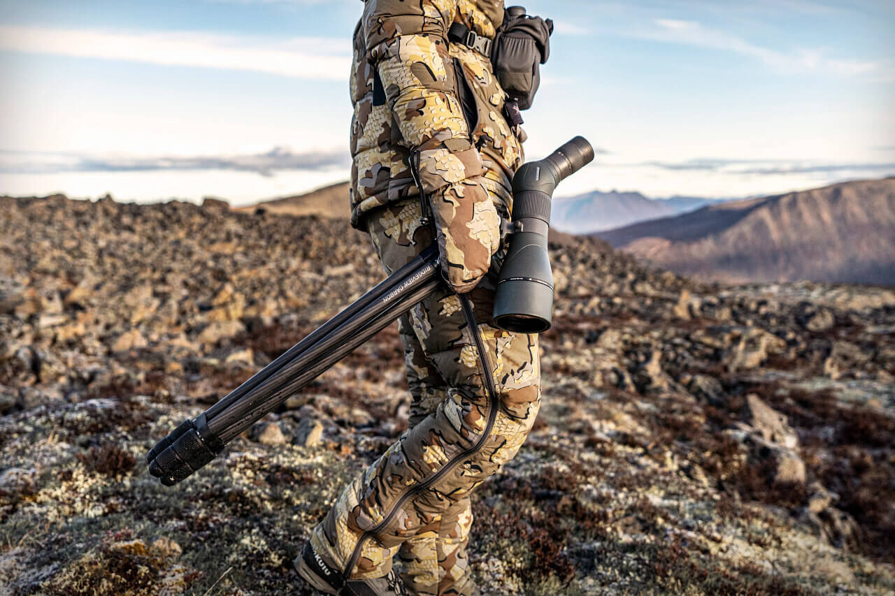 Individuum in Camouflage mit Fernglas und Stativ auf felsigem Gelände mit Bergen im Hintergrund