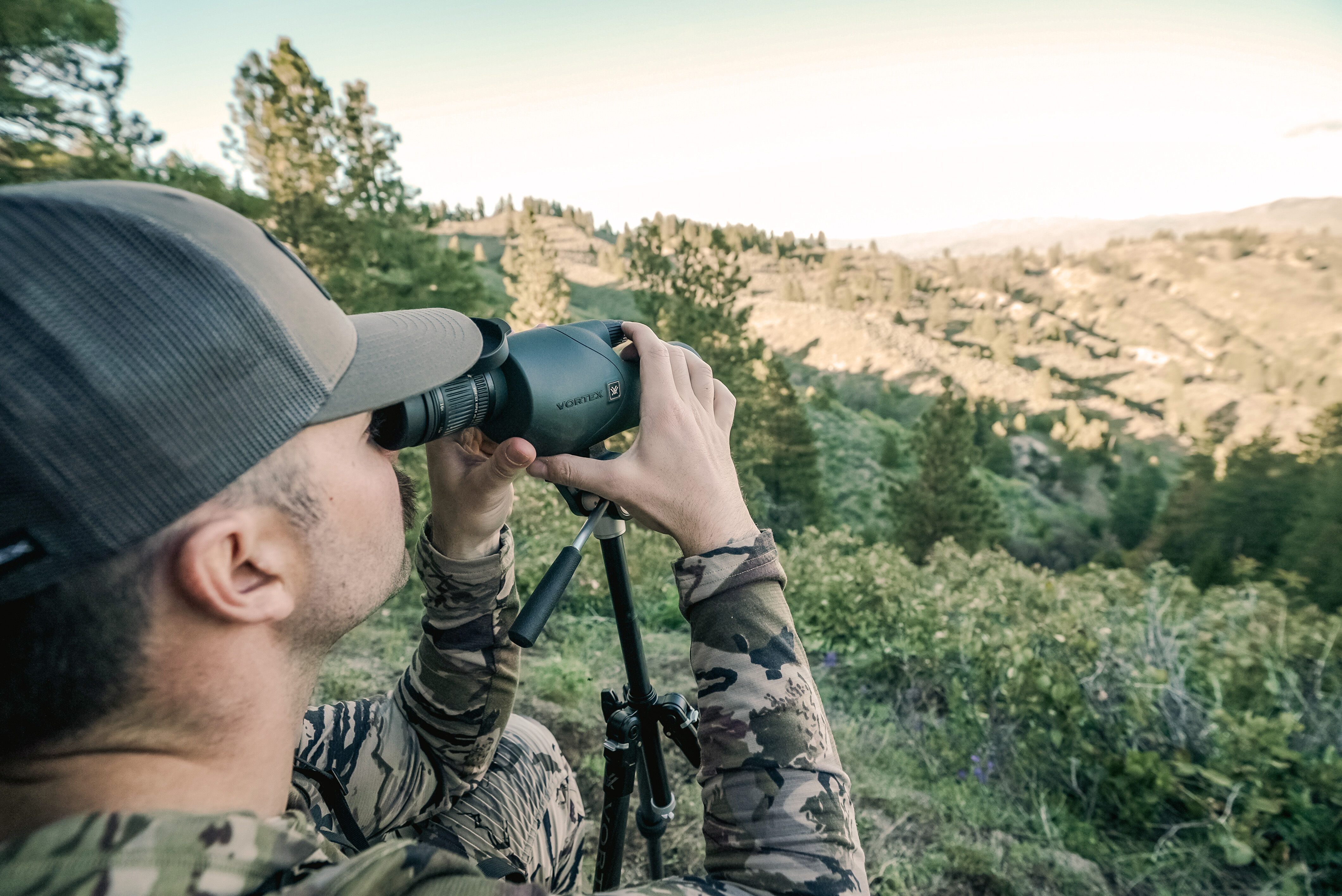 Mann im Tarnanzug beobachtet durch Fernglas in bergiger Landschaft. Person im Tarnanzug nutzt Fernglas auf einem Stativ, um eine bewaldete Berglandschaft zu beobachten.