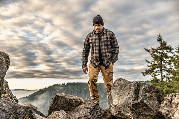 Mann mit Mütze und karierter Jacke steht auf einem felsigen Berg mit bewölktem Himmel im Hintergrund