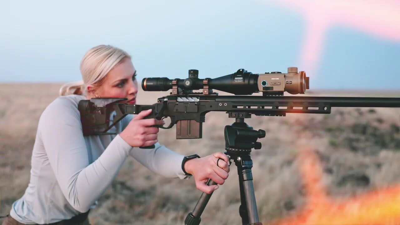Frau zielt mit Gewehr und montierter Optik auf einem Stativ in der Natur