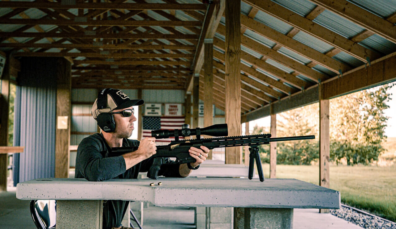 Ein Mann mit Gehörschutz und Sonnenbrille schießt mit einem Präzisionsgewehr auf einem überdachten Schießstand.