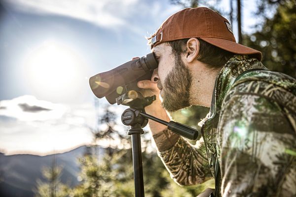 Ein Mann mit Camo-Jacke und Baseballkappe benutzt ein Fernglas auf einem Stativ in einer Waldlandschaft bei Sonnenuntergang.