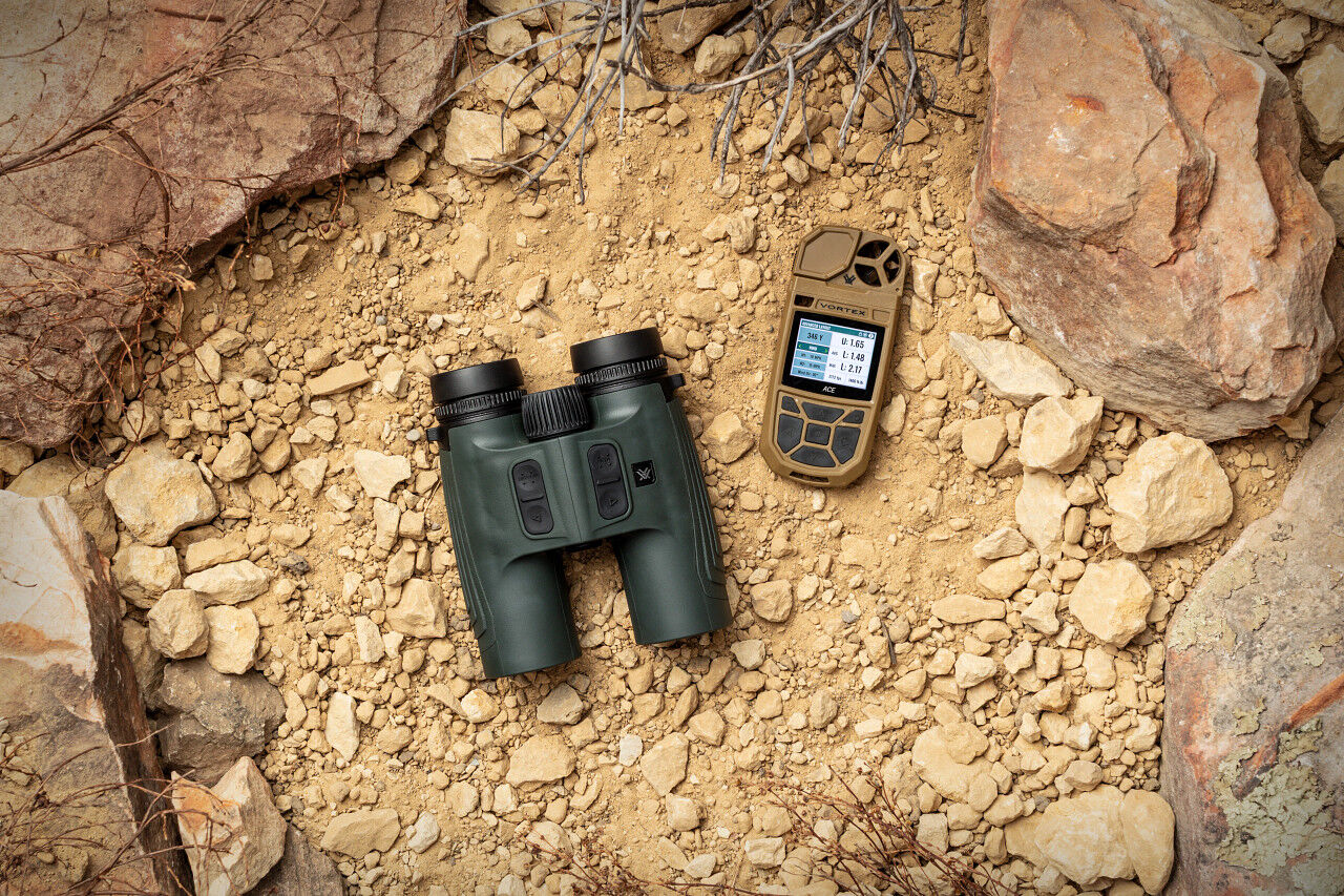 Grünes Outdoor-Fernglas mit integriertem Entfernungsmesser neben einem sandfarbenen taktischen Messgerät auf steinigem Wüstenboden