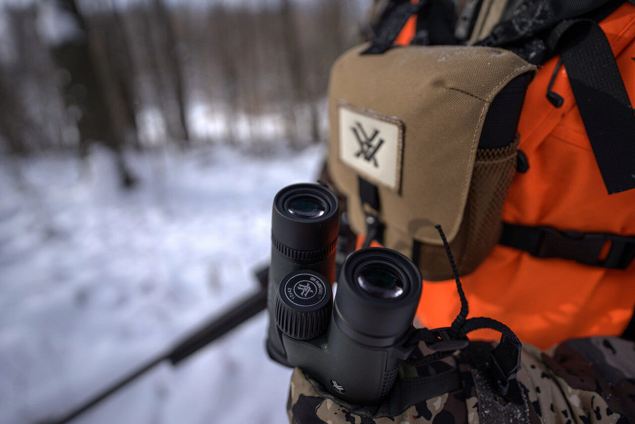 Nahaufnahme eines Jägers in Tarnkleidung im verschneiten Wald, der ein Fernglas hält, während er eine braune Ausrüstungstasche trägt.