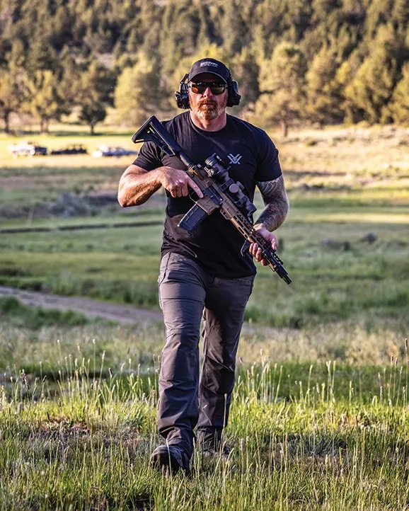 Mann beim Schießtraining im Freien, trägt taktische Outdoor-Hose und hält ein Gewehr mit Optik im offenen Gelände