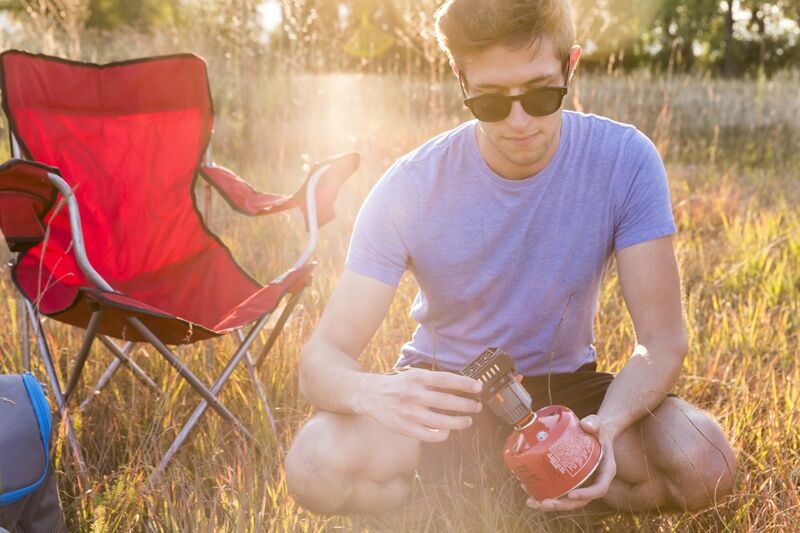 Junger Mann sitzt beim Campen auf einer Wiese und montiert einen kompakten Gaskocher auf eine rote Gaskartusche neben einem Campingstuhl
