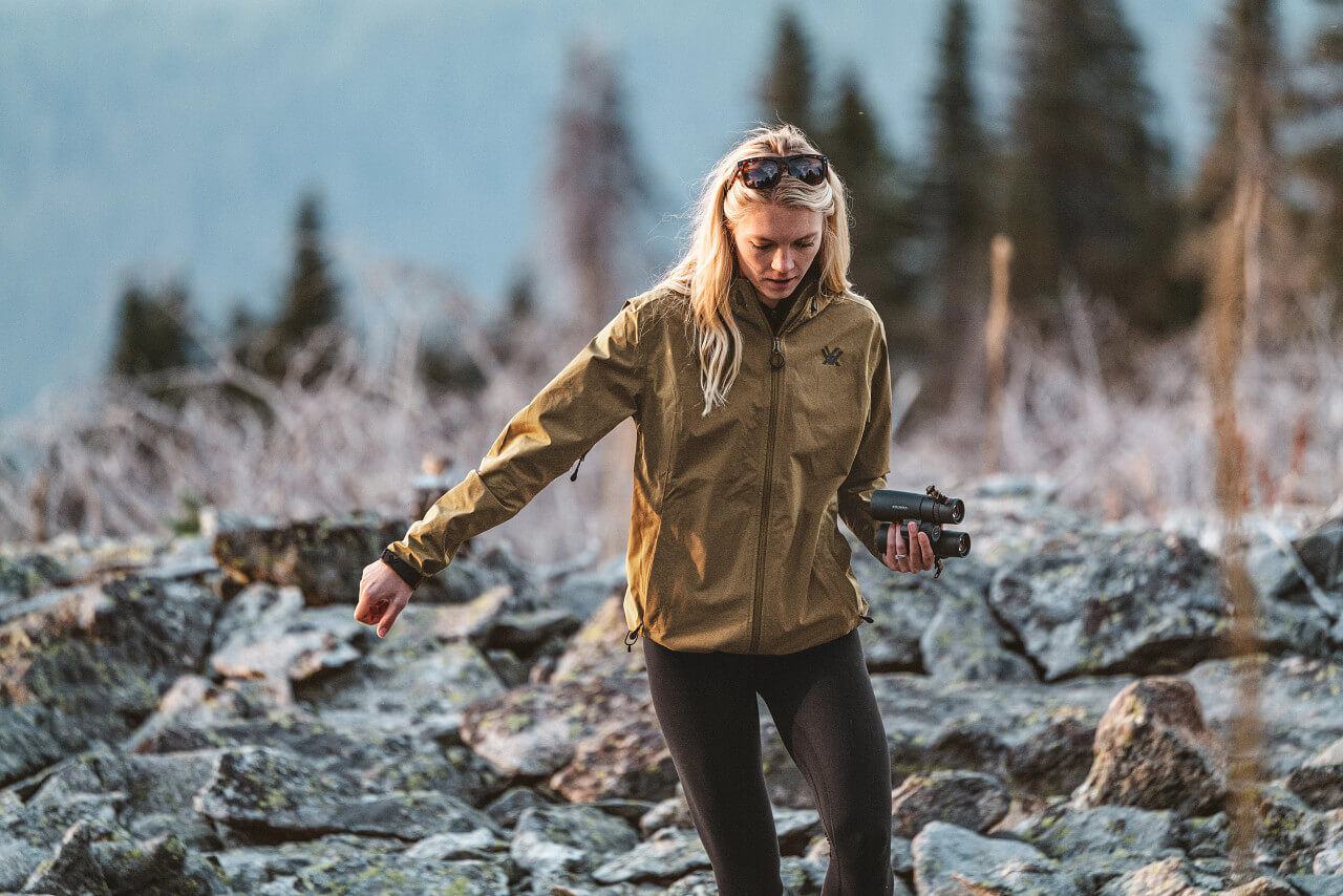 Eine Frau mit blonder Haaren trägt eine braune Outdoor-Jacke und hält ein Fernglas in der Hand, während sie auf einem felsigen Pfad in einer bewaldeten Landschaft wandert.