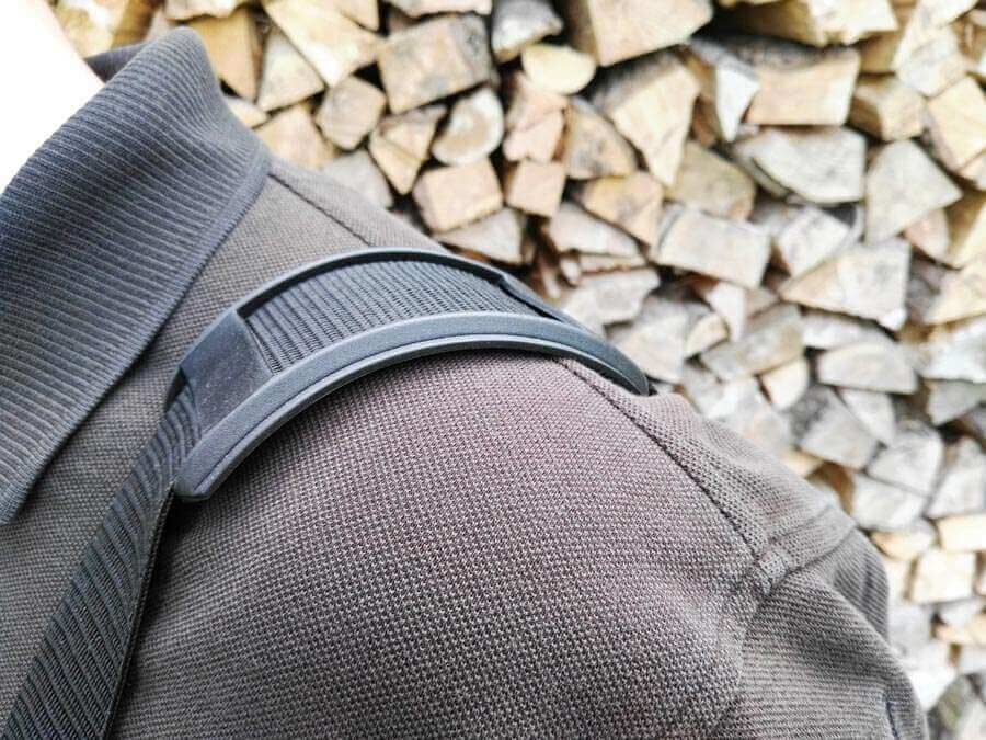 Ein schwarzer Schultergurt eines Rucksacks auf einer dunkelbraunen Jacke vor einem Hintergrund aus gestapeltem Brennholz