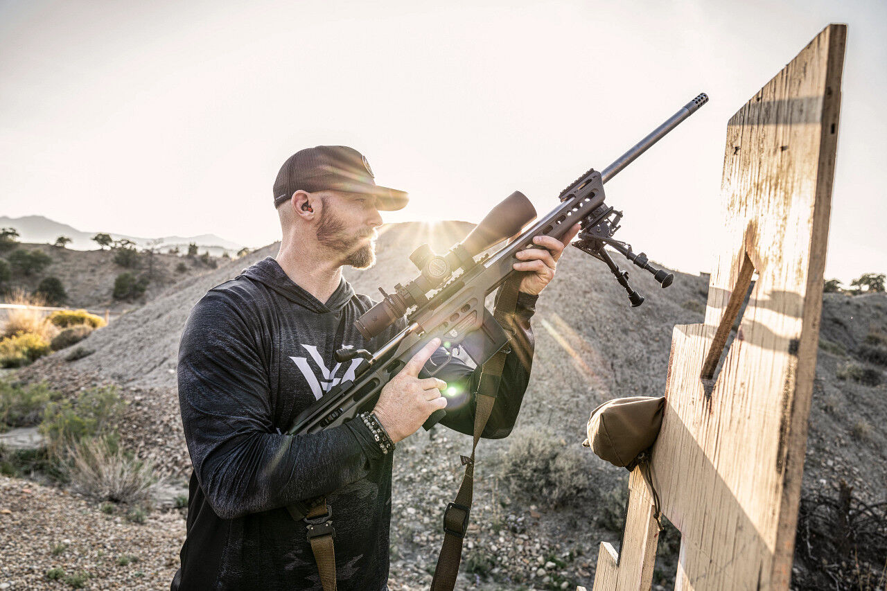 Ein Mann in sportlicher Kleidung hält ein Präzisionsgewehr und zielt durch ein Zielfernrohr, während er im Freien steht. Im Hintergrund sind hügelige Landschaften sichtbar.