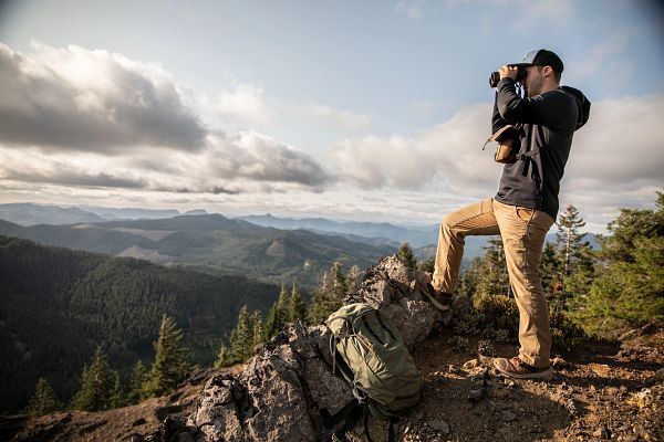 Ein Wanderer steht auf einem Felsvorsprung und schaut durch ein Fernglas in die Ferne, umgeben von bewaldeten Bergen und Wolken am Himmel.