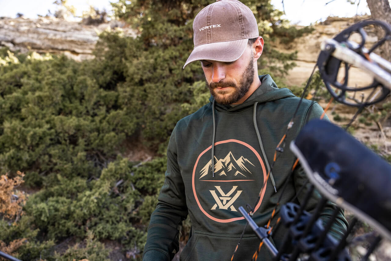 Mann trägt eine Vortex-Kappe und einen grünen Hoodie mit Bergmotiv, während er draußen im Wald mit einem Bogen hantiert