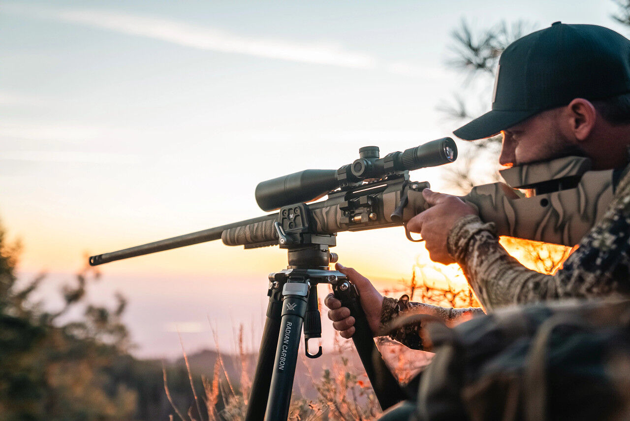 Ein Jäger zielt mit einem Gewehr, das auf einem Dreibein-Stativ befestigt ist, während die Sonne am Horizont untergeht.
