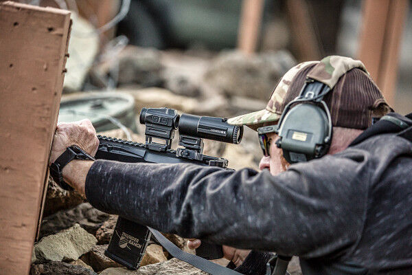Mann mit Tarnkappe und Gehörschutz zielt mit einem Gewehr in einer Trainingsumgebung im Freien