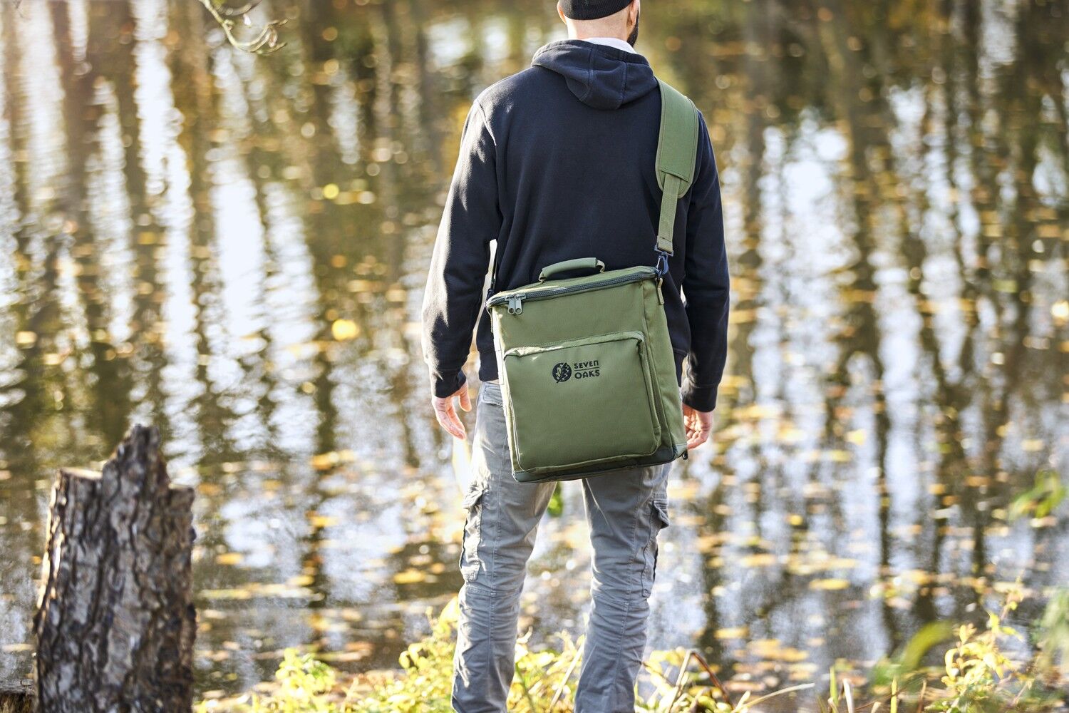 Rückansicht eines Mannes in schwarzer Kapuzenjacke mit einer grünen Seven Oaks Schultertasche am Flussufer