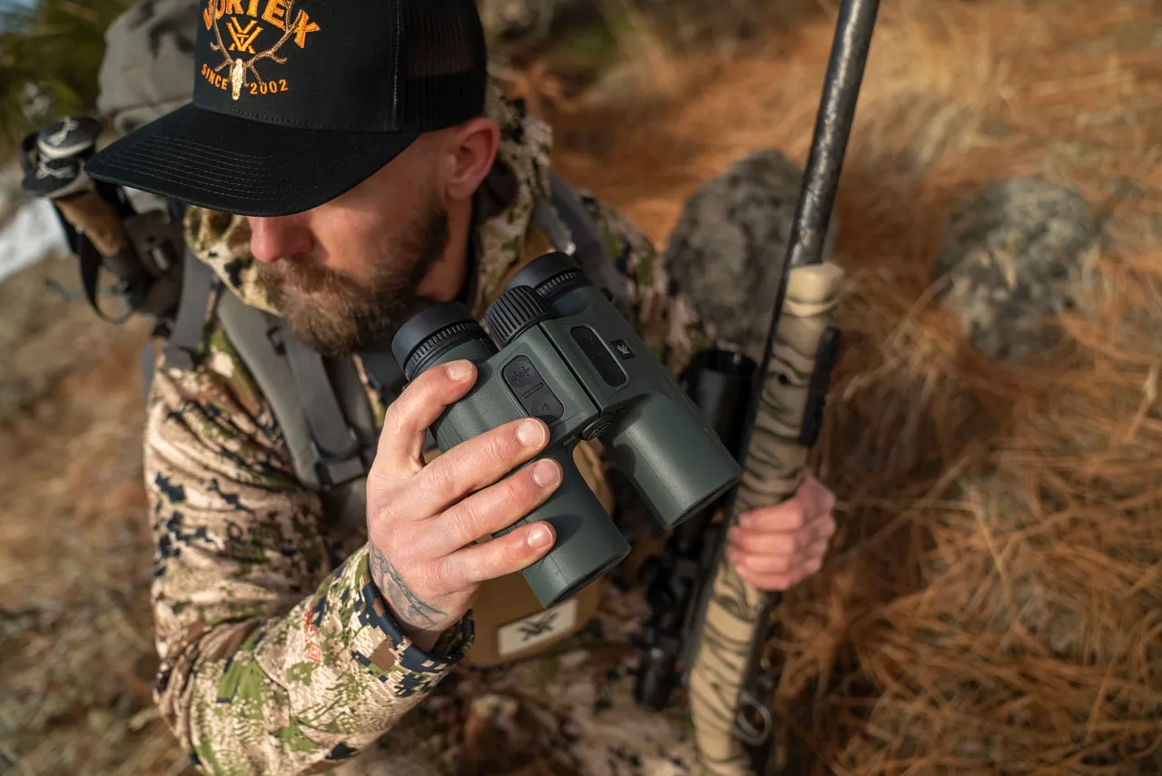Jäger in Tarnkleidung hält ein robustes, grünes Jagdfernglas in der Hand und trägt ein Jagdgewehr im herbstlichen Gelände